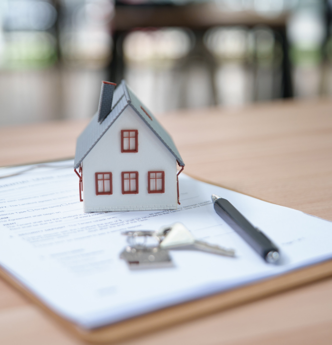 A model of a house sits on a clipboard with a paper application, next to a pen and set of keys